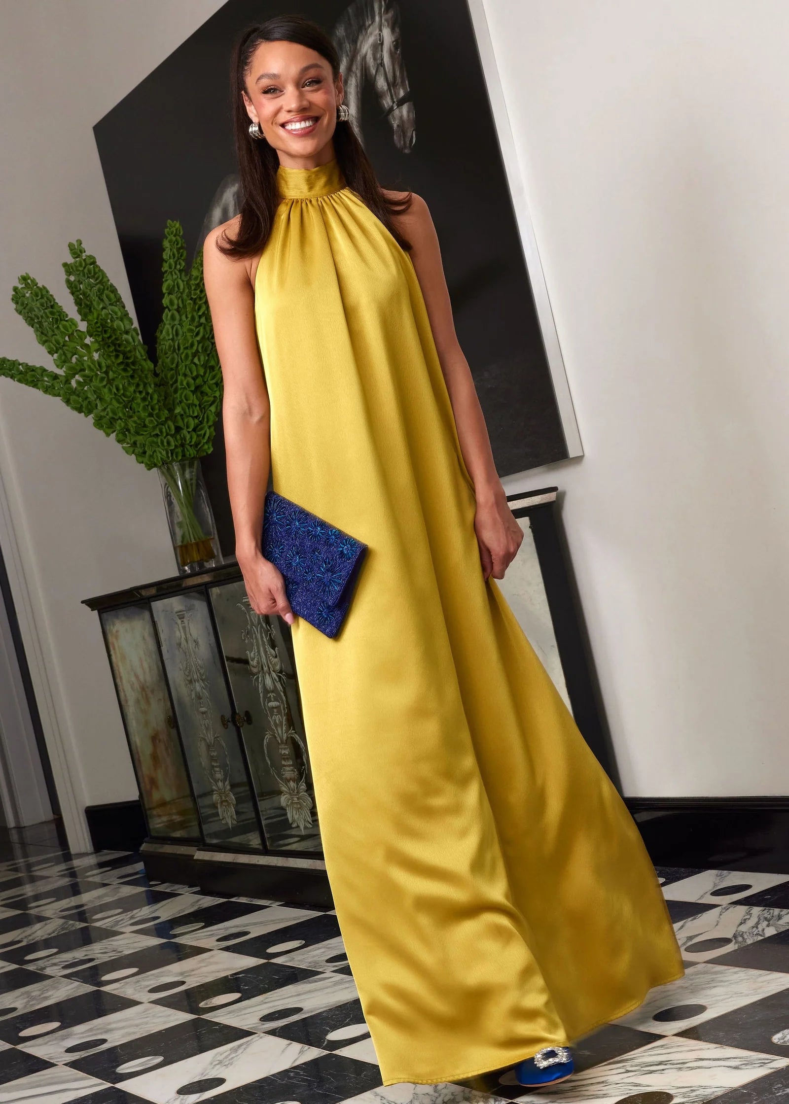A woman stands elegantly in a full-length golden olive satin gown with a high neckline and flowing silhouette, holding a blue clutch.