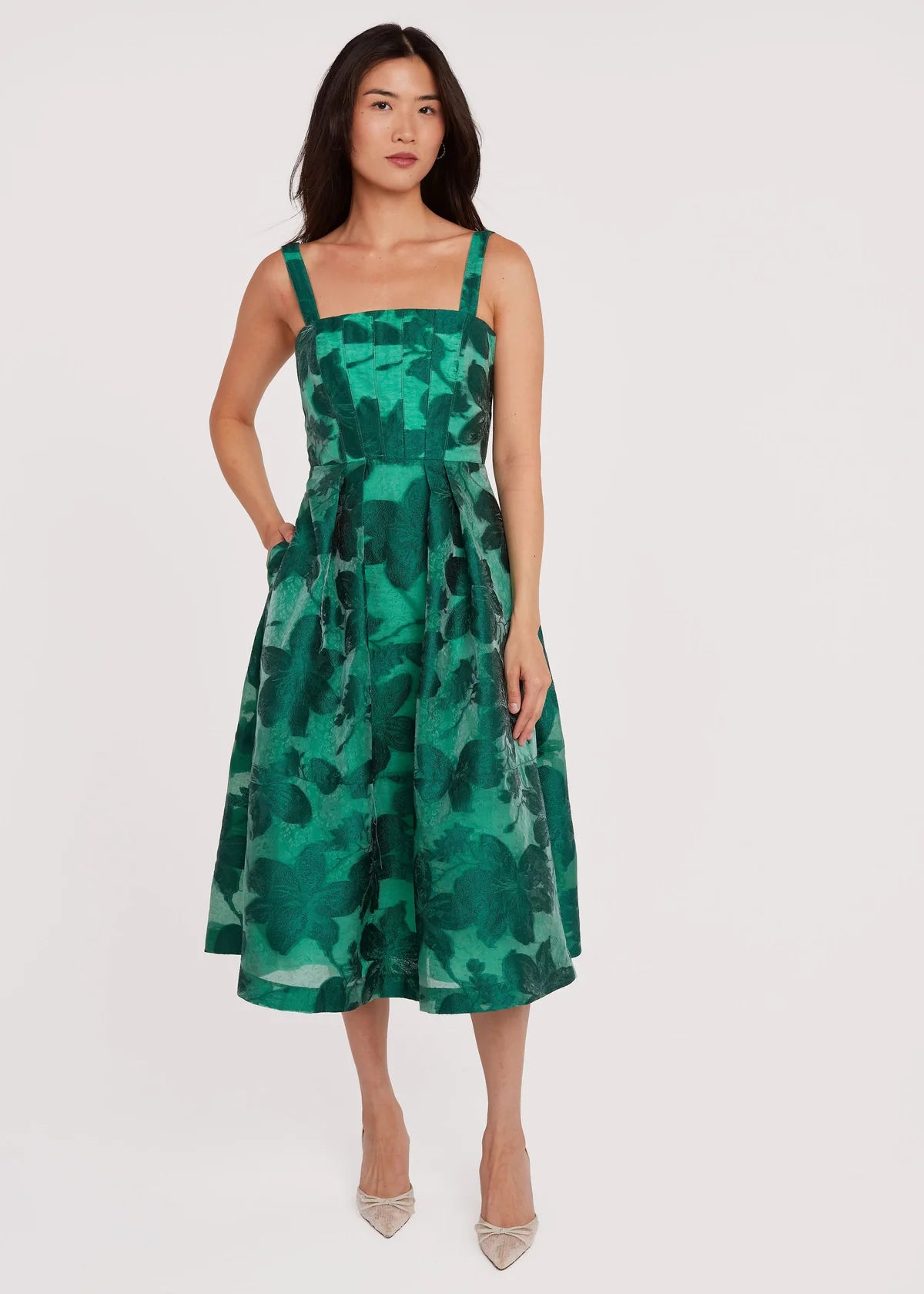 A model showcases a vibrant green dress with a structured bodice and spaghetti straps, featuring a flared skirt and pockets.