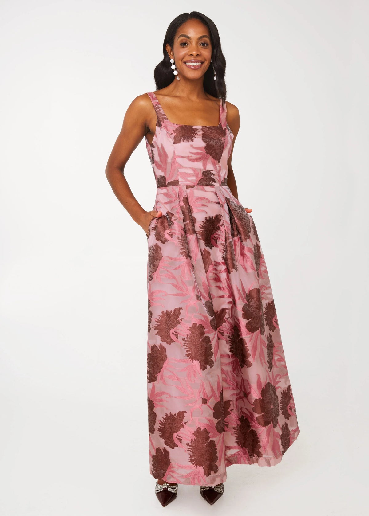 A woman stands confidently in a blush pink gown adorned with dark floral patterns, featuring a square neckline and pockets.