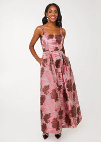 A woman stands confidently in a blush pink gown adorned with dark floral patterns, featuring a square neckline and pockets.