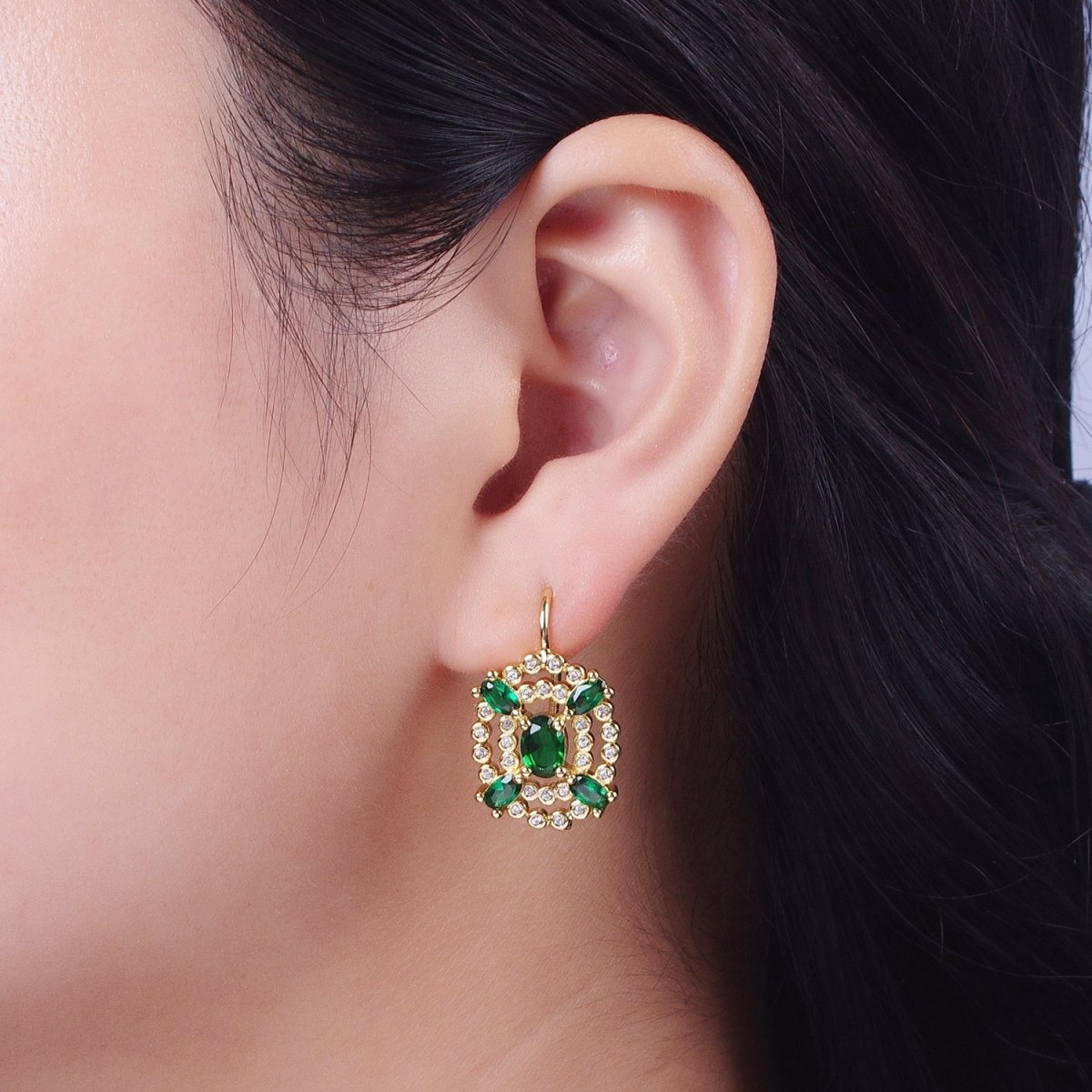 A close-up of a womans ear showcasing a beautiful gold circular drop earring adorned with green gemstones and small clear stones. This elegant accessory is perfect for special occasions like galas or black tie weddings.