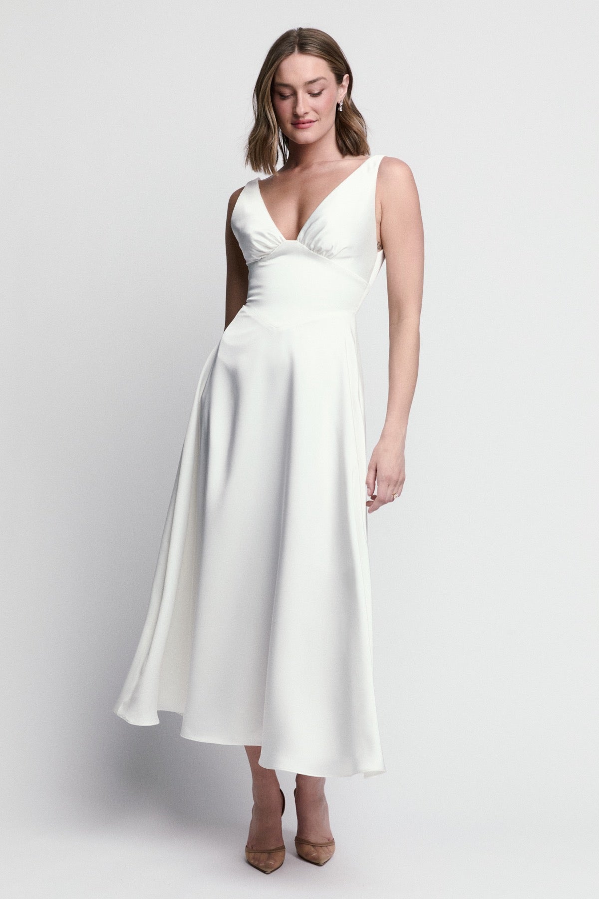 A model poses gracefully in a sleeveless, white midi dress with a deep V-neckline, highlighting a flattering silhouette.