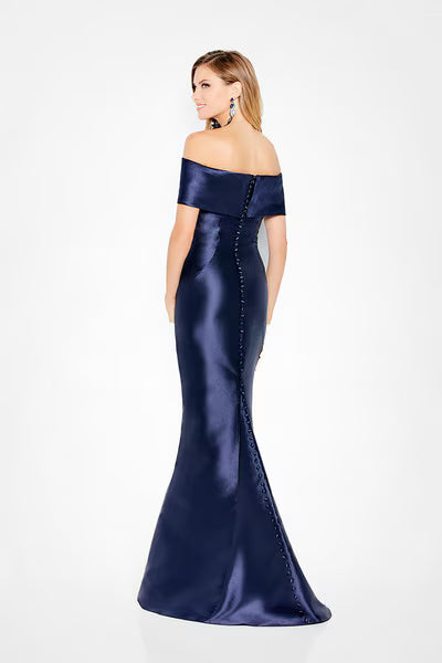 A woman stands in profile wearing a stunning navy off-the-shoulder mikado trumpet gown, showcasing a pleated V-neckline and elegant fold-over back detail with covered buttons.