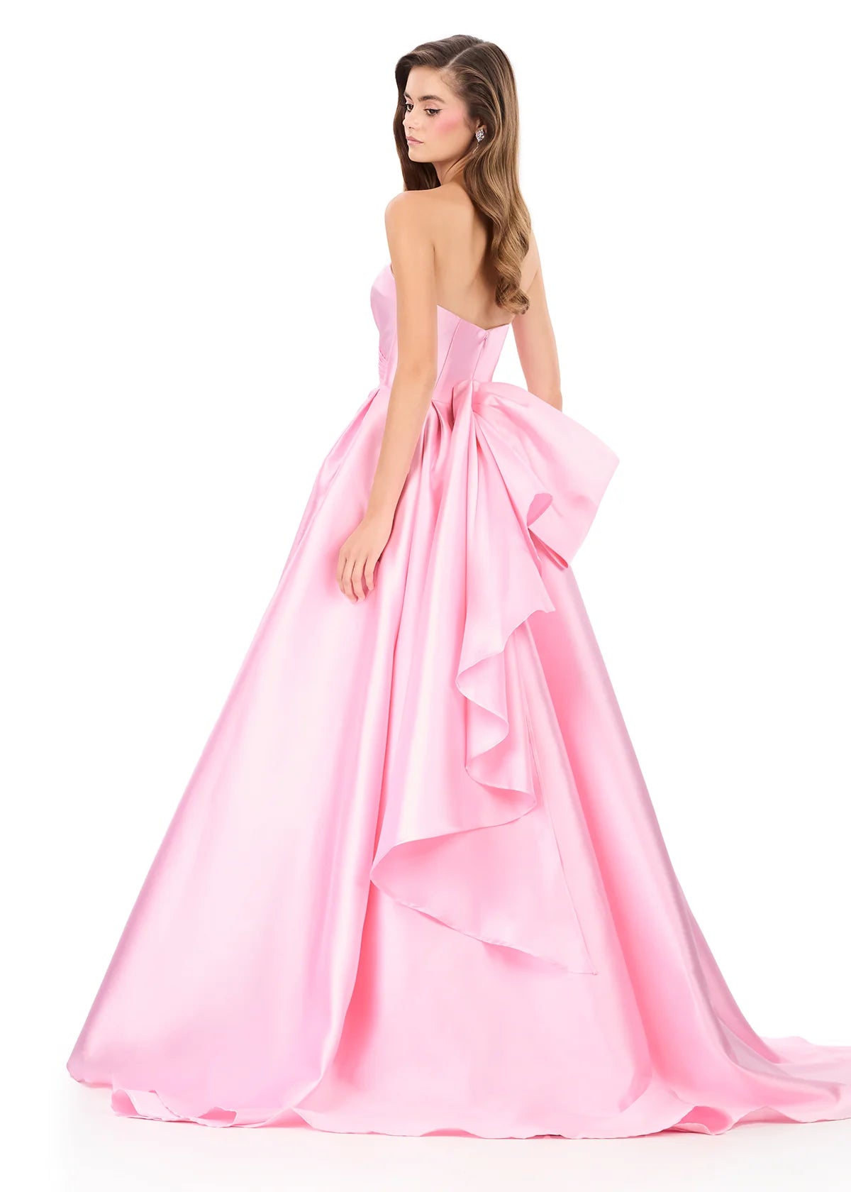A model showcases a stunning, strapless ice pink gown featuring a voluminous skirt and a large bow at the back, perfect for formal events like proms, galas, or weddings. This elegant dress is available near Apex, Cary, and Raleigh.