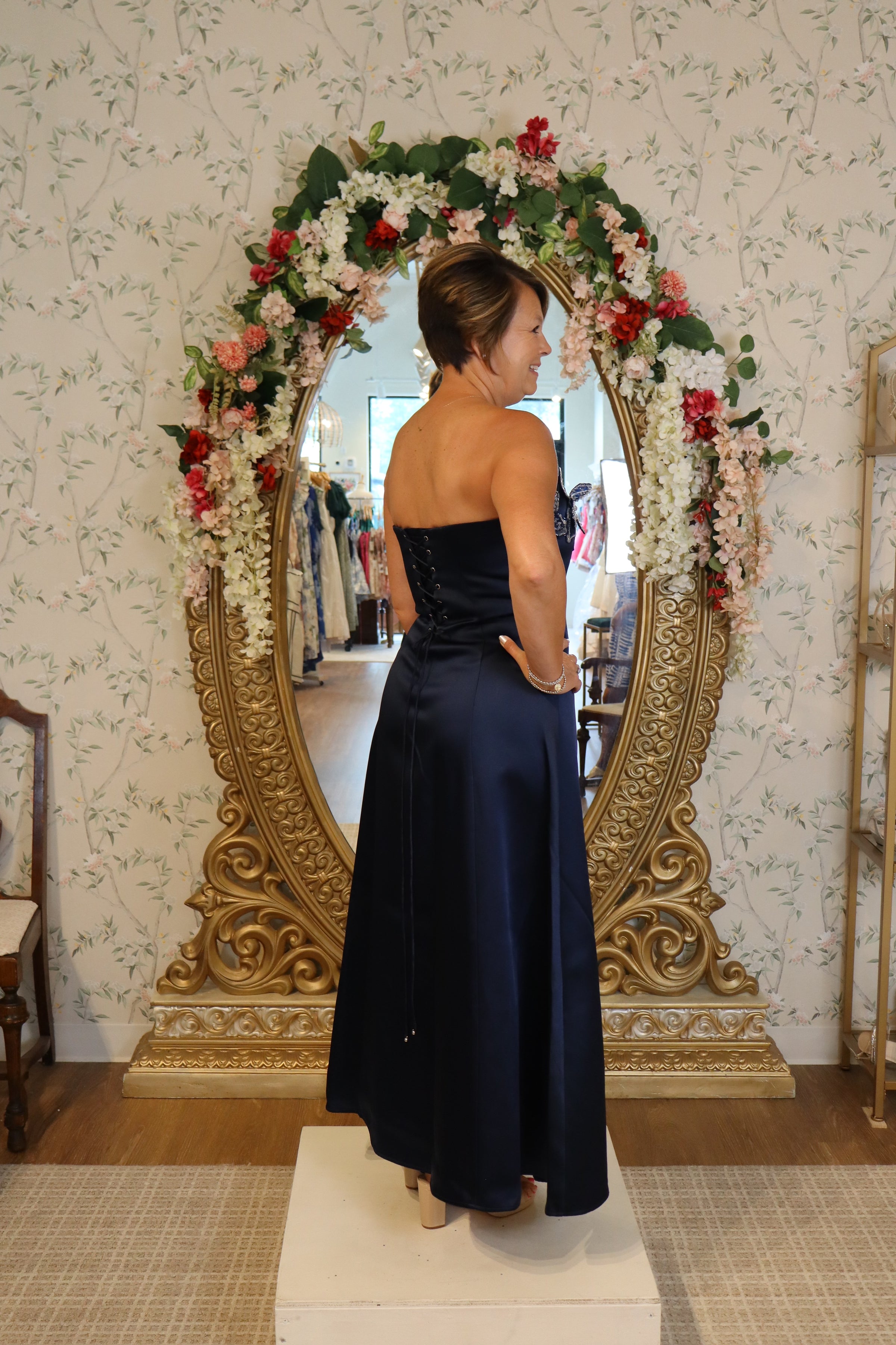 A woman is modeling a navy fit and flare gown with a strapless design, showcasing its elegant back lacing. This gown is perfect for occasions like a mother of the bride, prom, or a gala, and is available near Apex, Cary, and Raleigh.