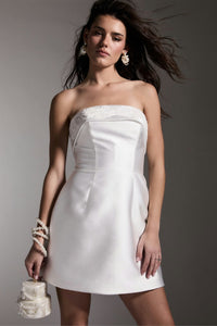 A model showcases a strapless white dress featuring a lace detail along the neckline, styled with a pearl bracelet and holding a small handbag.