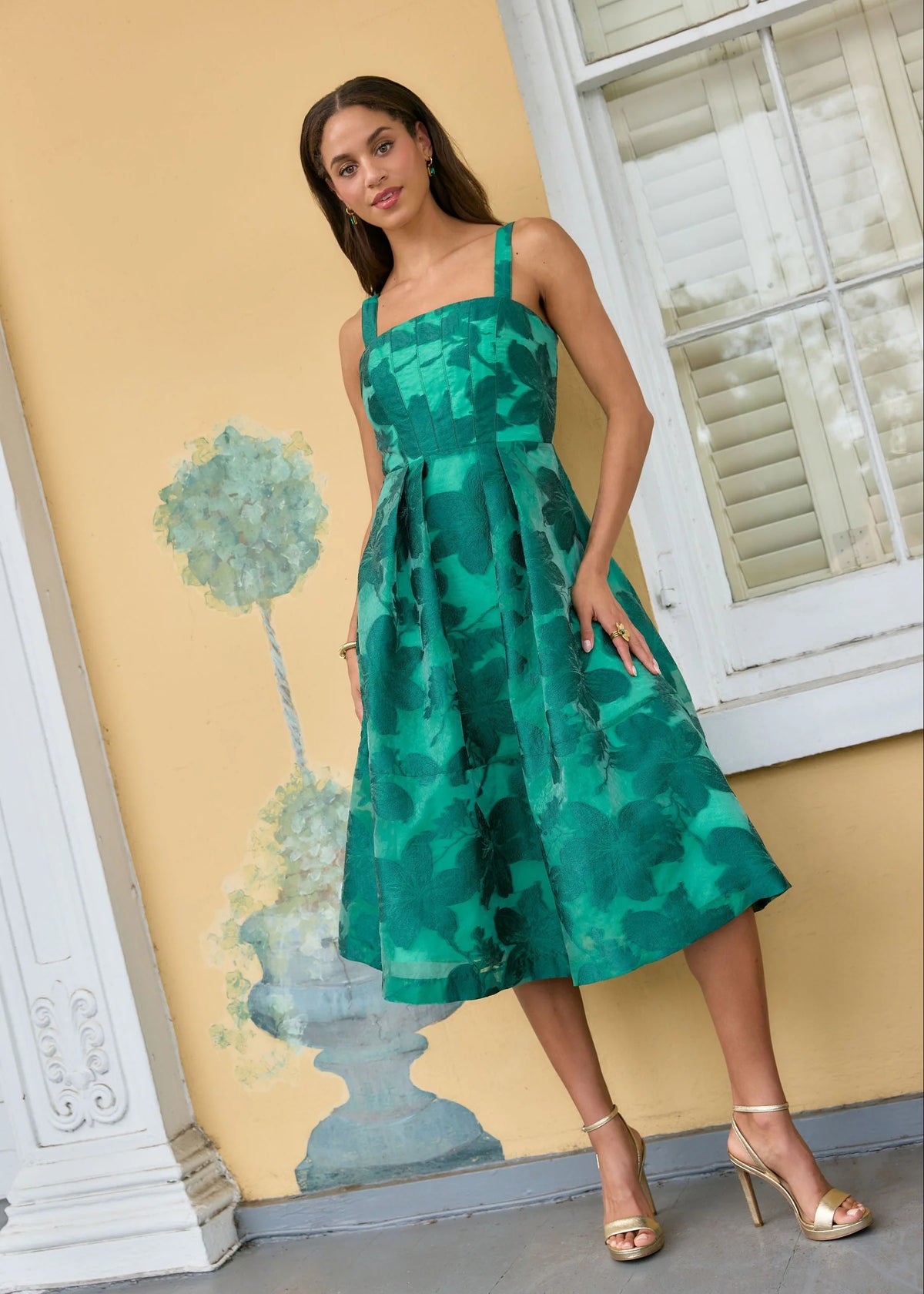 A model stands in front of a pastel yellow wall adorned with a decorative plant painting, wearing a vibrant green floral dress with a structured bodice and wide straps.