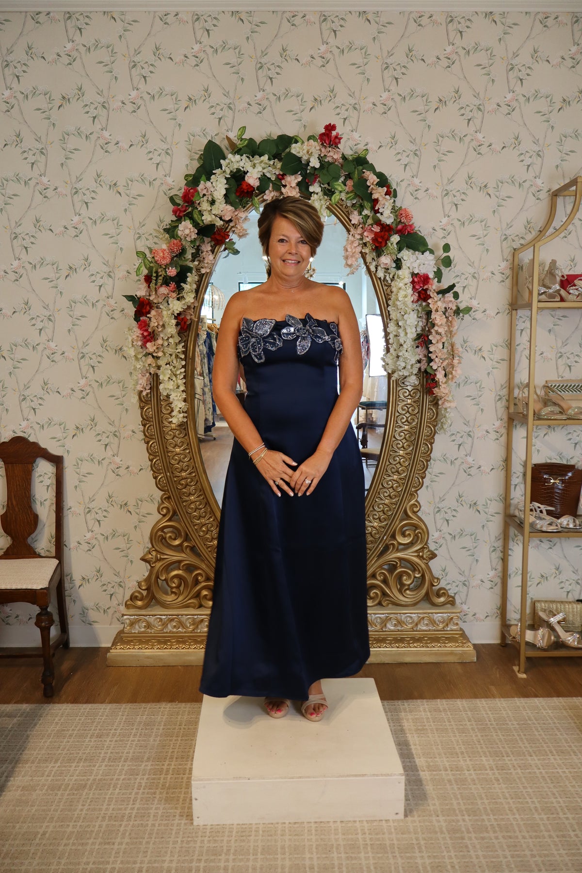 A woman stands confidently in a navy fit and flare gown with intricate beading, posing in front of a large ornate mirror adorned with flowers.