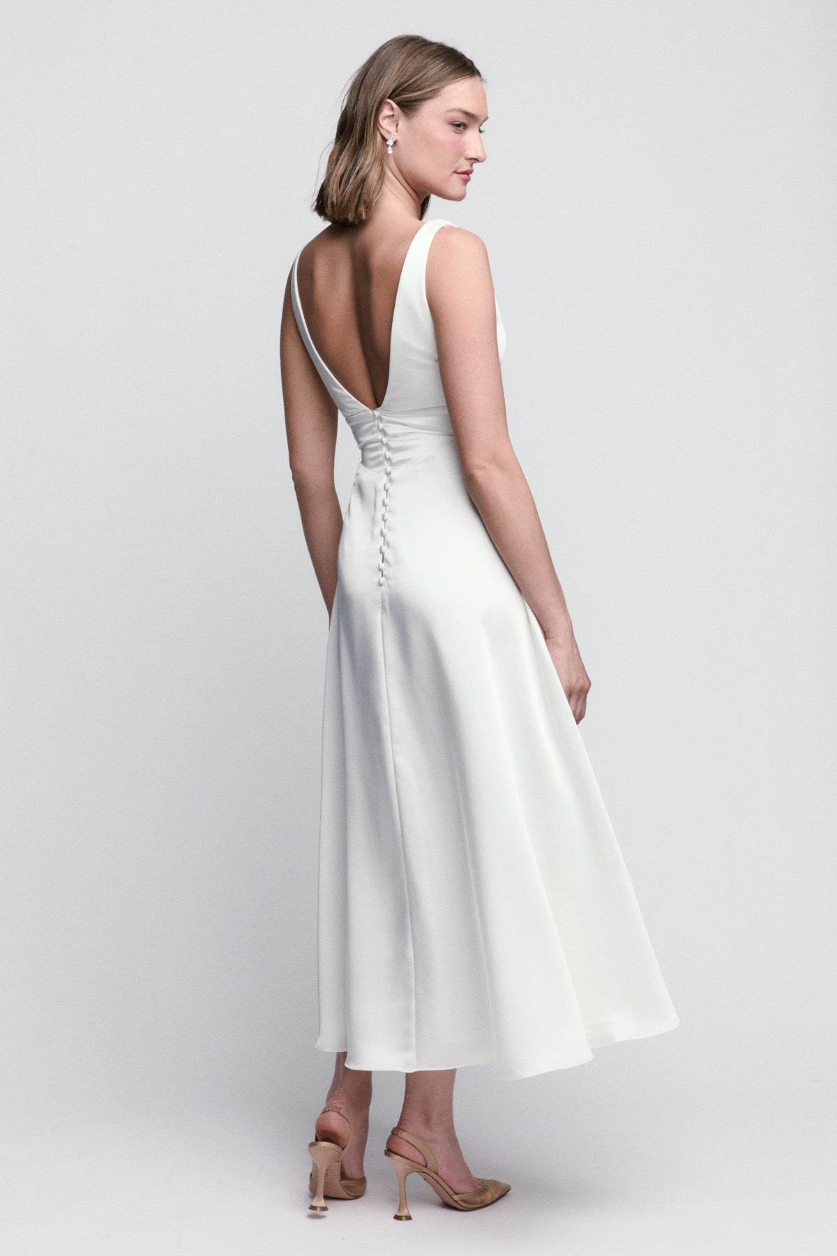 A model showcases a chic, sleeveless white dress with a deep V-back and a flowing midi-length skirt. This elegant gown is perfect for special occasions like weddings or formal events, and is conveniently available near Apex, Cary,.
