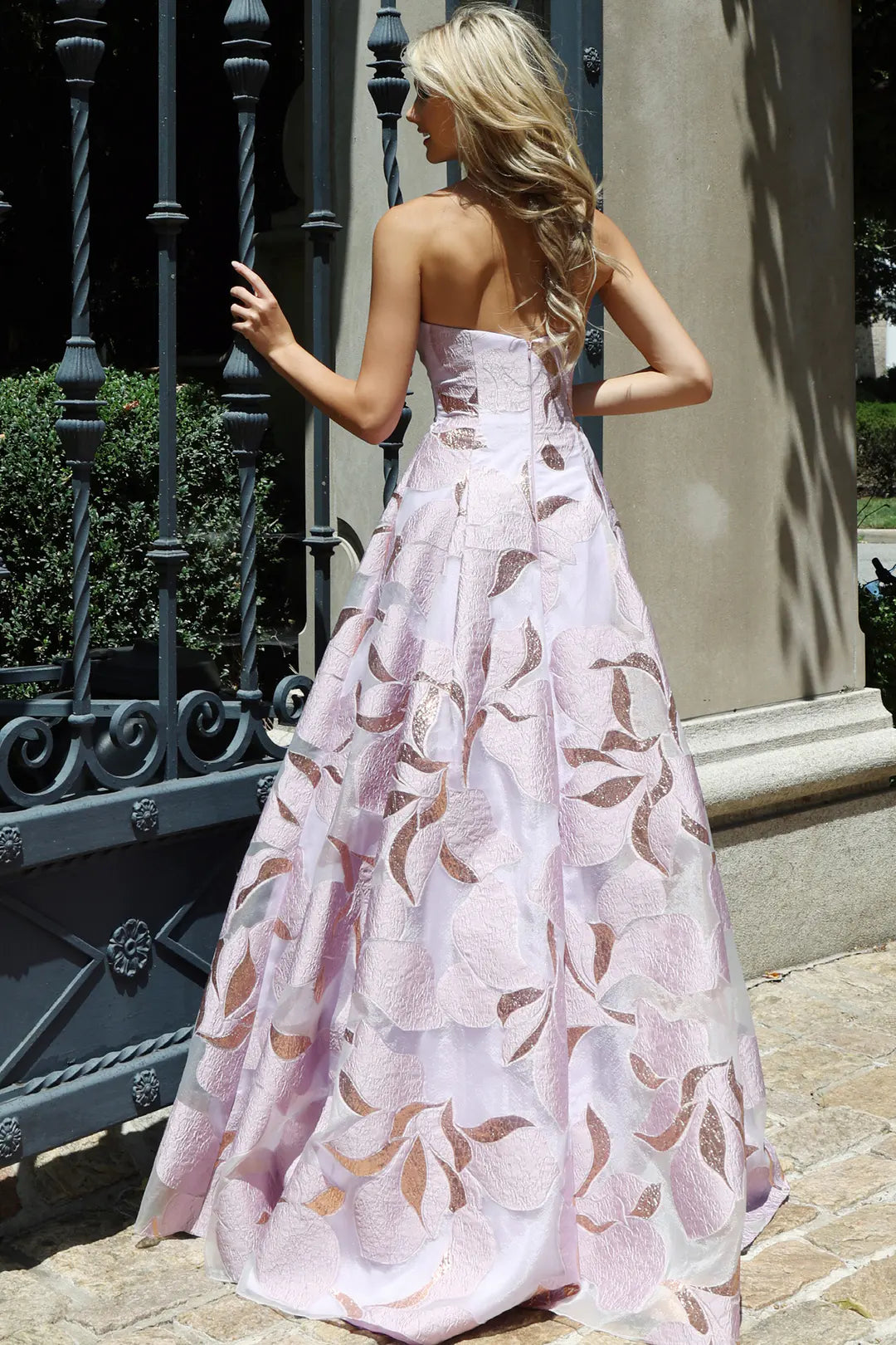 A model showcases a stunning lilac gown with intricate floral patterns, standing gracefully with her back to the camera.