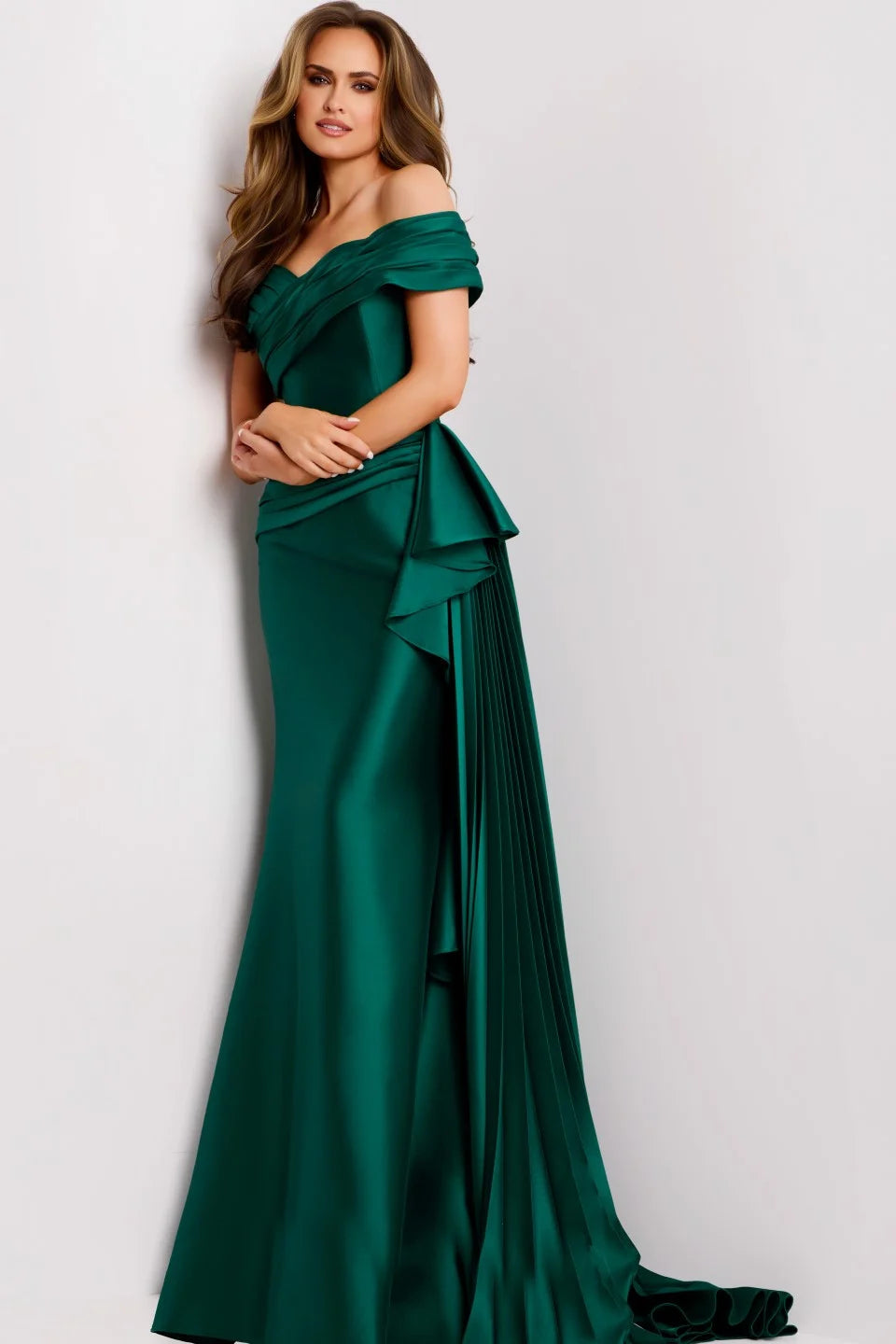 A model poses in a stunning emerald green floor-length gown with an off-the-shoulder sweetheart neckline and soft pleats cascading down one side.