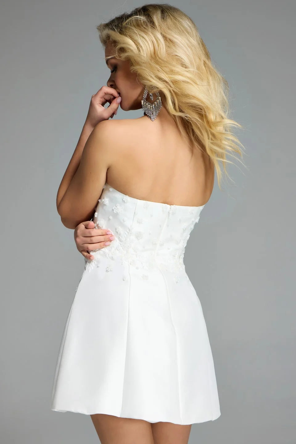 A model poses in a strapless off-white dress with intricate floral detailing on the back, showcasing a fitted bodice and a flared skirt.