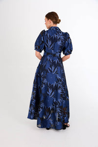 A woman is seen from the back, wearing a long, elegant gown with a blue floral pattern on a dark background. This stunning dress is perfect for special occasions such as galas or weddings, conveniently located near Apex, Cary, and Raleigh.