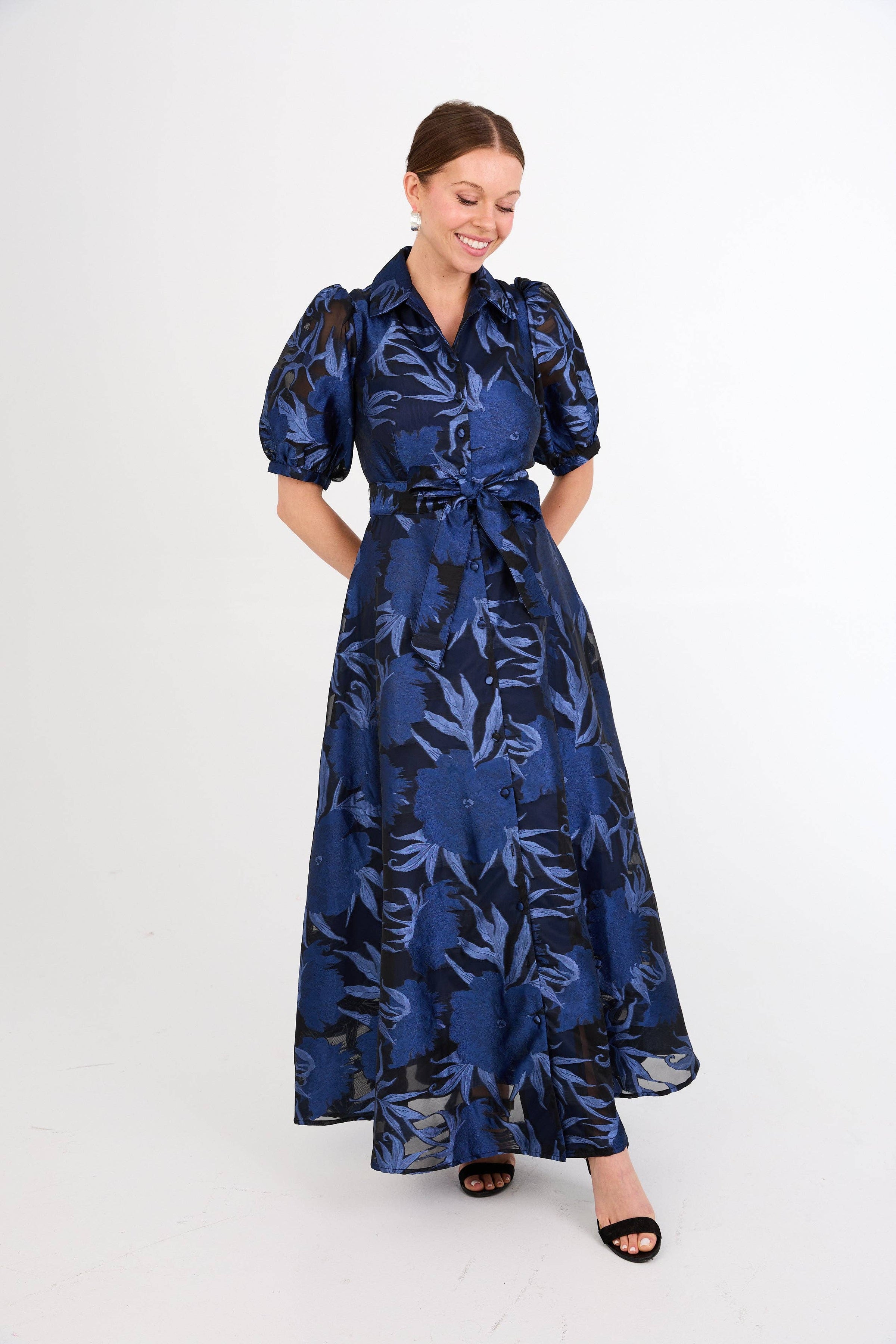 A woman is wearing a stunning royal blue gown with a floral pattern, featuring puff sleeves and a belted waist.