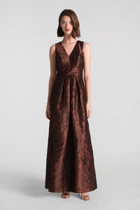 A model poses in a floor-length chocolate brown gown with a textured jacquard fabric and a v-neckline, perfect for elegant occasions like galas or weddings. This stunning dress is conveniently located near Apex, Cary, and Raleigh.
