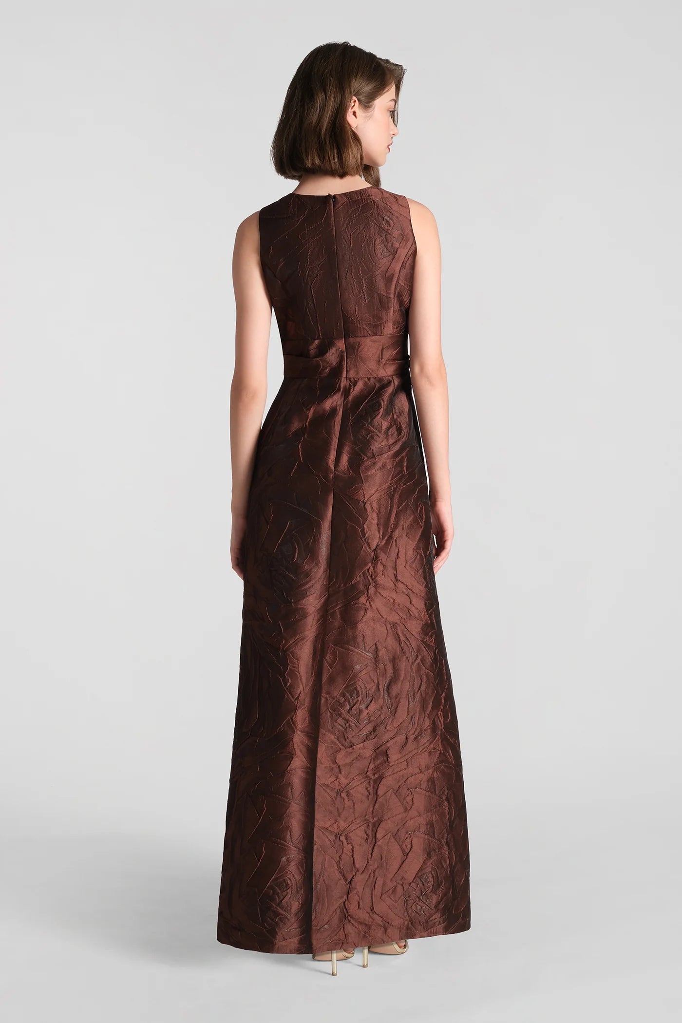 A model is seen from the back wearing a stunning chocolate brown gown made from textured jacquard fabric. This elegant dress is perfect for formal occasions such as galas, weddings, or proms and is available near Apex, Cary, and Raleigh.