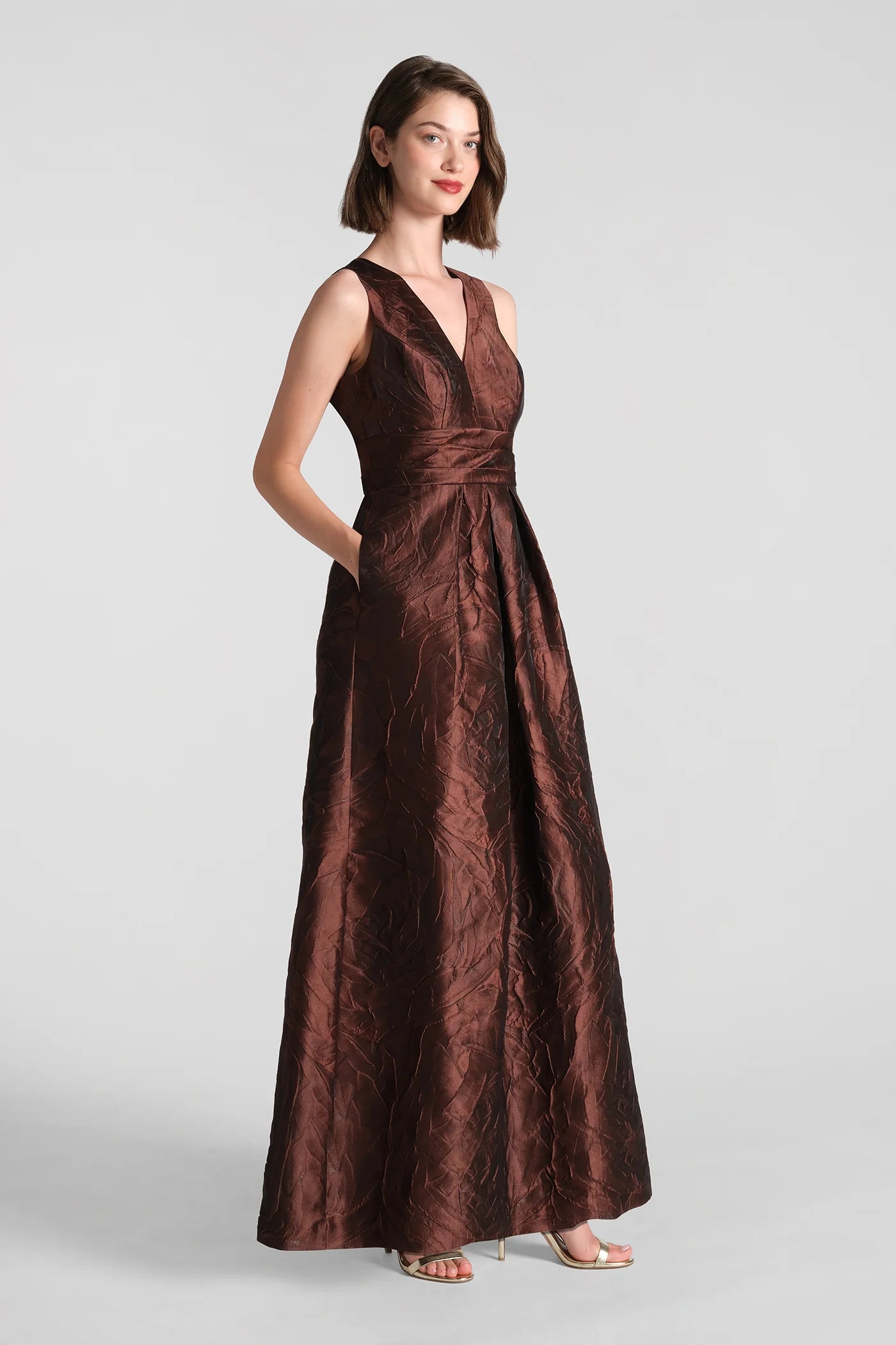 A model showcases the elegant Mariah Gown in rich chocolate brown, featuring a textured jacquard fabric and a sophisticated silhouette with pockets.