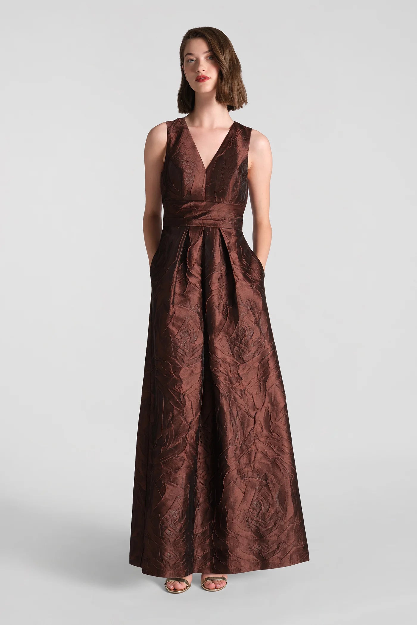 A model showcases the elegant Mariah Gown in rich chocolate brown, featuring a deep V-neckline and a flowing skirt with pockets.