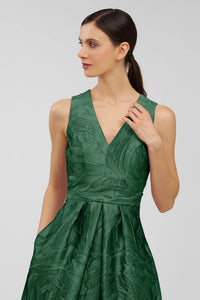 A woman models a stunning emerald green gown with a textured fabric and a V-neckline, featuring elegant details and pockets.