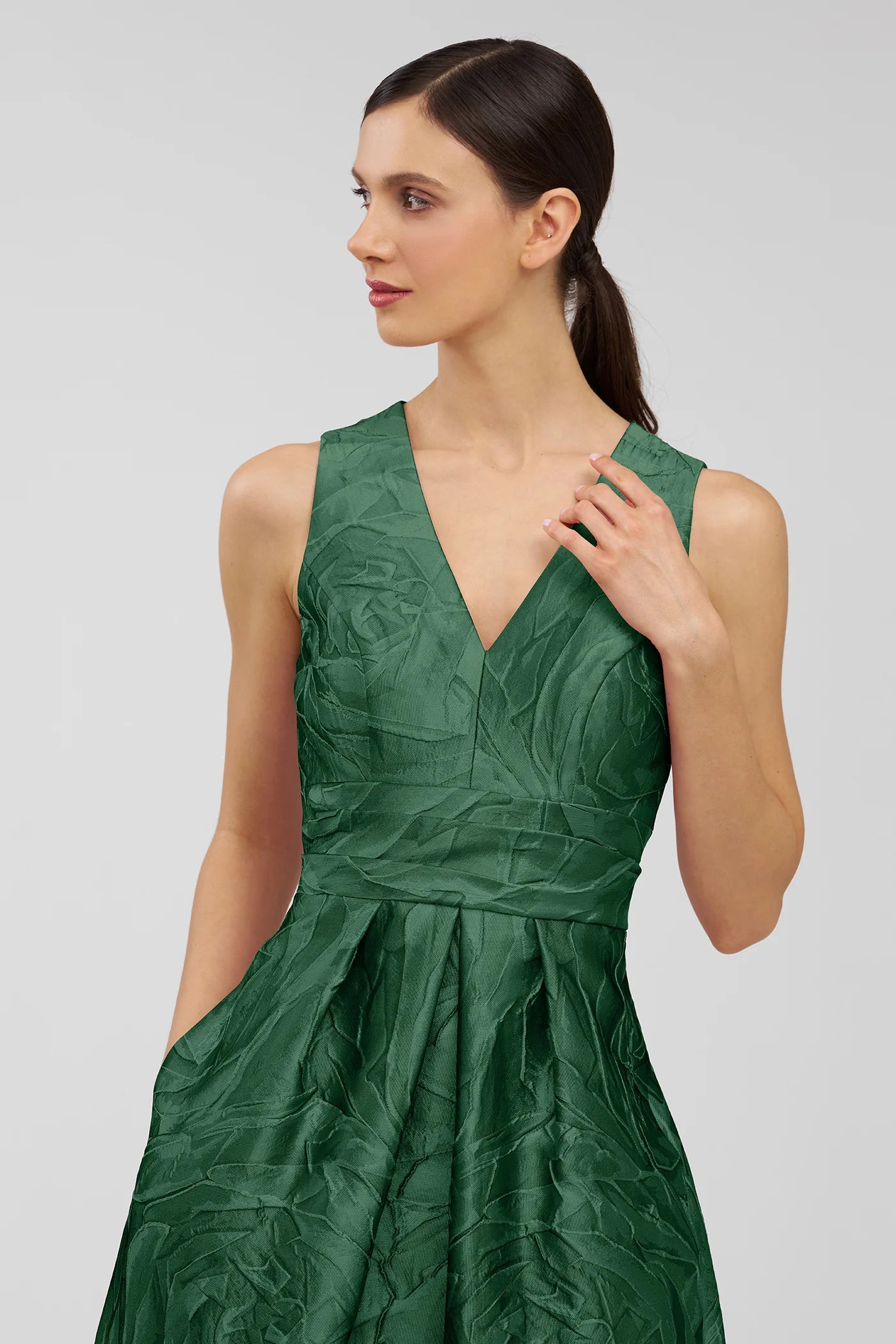 A woman models a stunning emerald green gown with a textured fabric and a V-neckline, featuring elegant details and pockets.