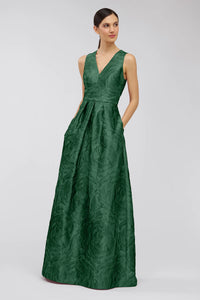 A model showcases the Mariah Gown in a rich emerald green, featuring a deep V-neckline and elegant floral jacquard texture.