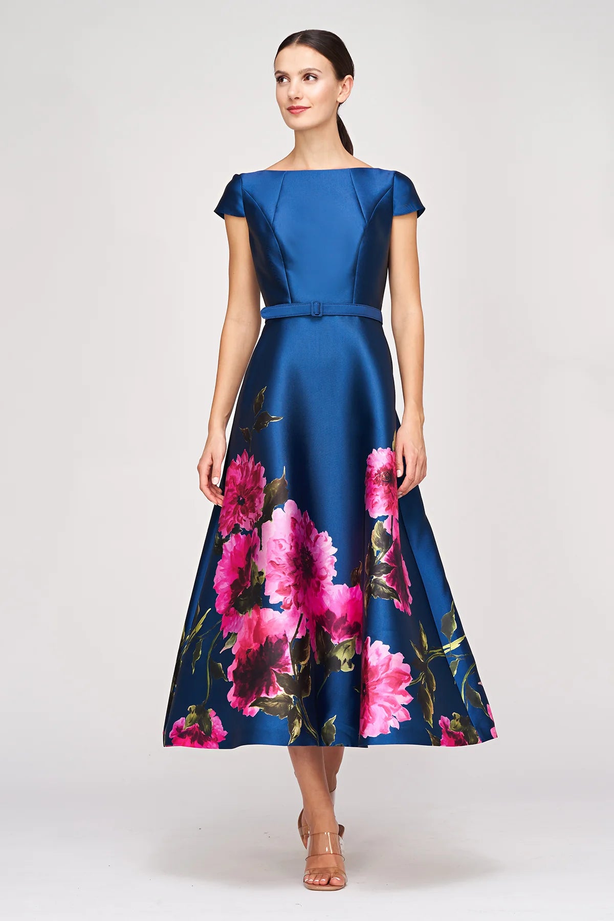 A model showcases a vibrant blue gown featuring large pink floral patterns along the hem, designed with short sleeves and a fitted bodice.