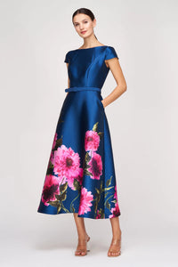 A woman poses in a stunning, knee-length dress featuring a fitted bodice and short sleeves. The dress is a rich blue satin adorned with vibrant pink floral patterns, perfect for occasions like weddings, galas, or formal events, and is.