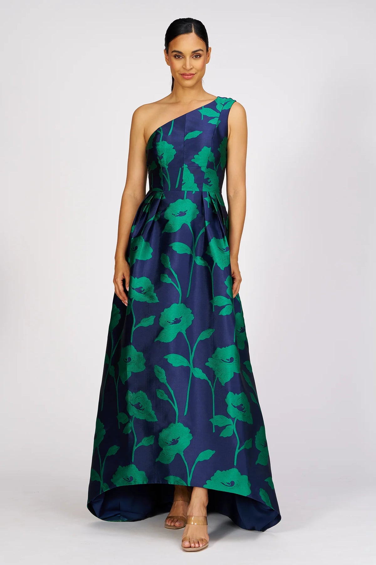 A model showcases a striking one-shoulder gown featuring oversized green floral patterns against a navy background. This elegant dress, perfect for a black tie wedding or gala, is conveniently available near Apex, Cary, and Raleigh.