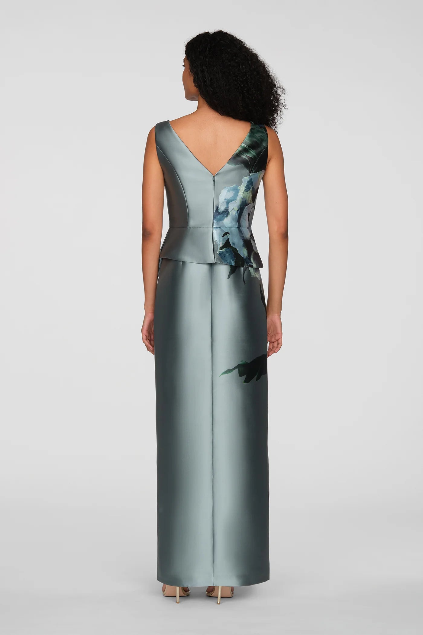 A woman is seen from the back wearing a sleek, floor-length gown with a V-neck and an asymmetrical peplum featuring a floral print.