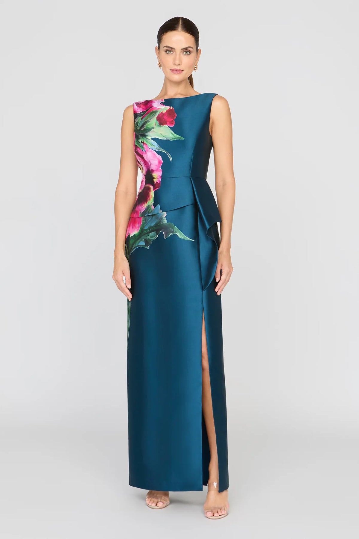 A model showcases a sleek blue gown adorned with vibrant floral prints, featuring a bateau neckline and an asymmetrical peplum with a ruffle.