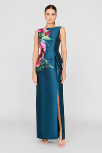 A model showcases a sleek blue gown adorned with vibrant floral prints, featuring a bateau neckline and an asymmetrical peplum with a ruffle.