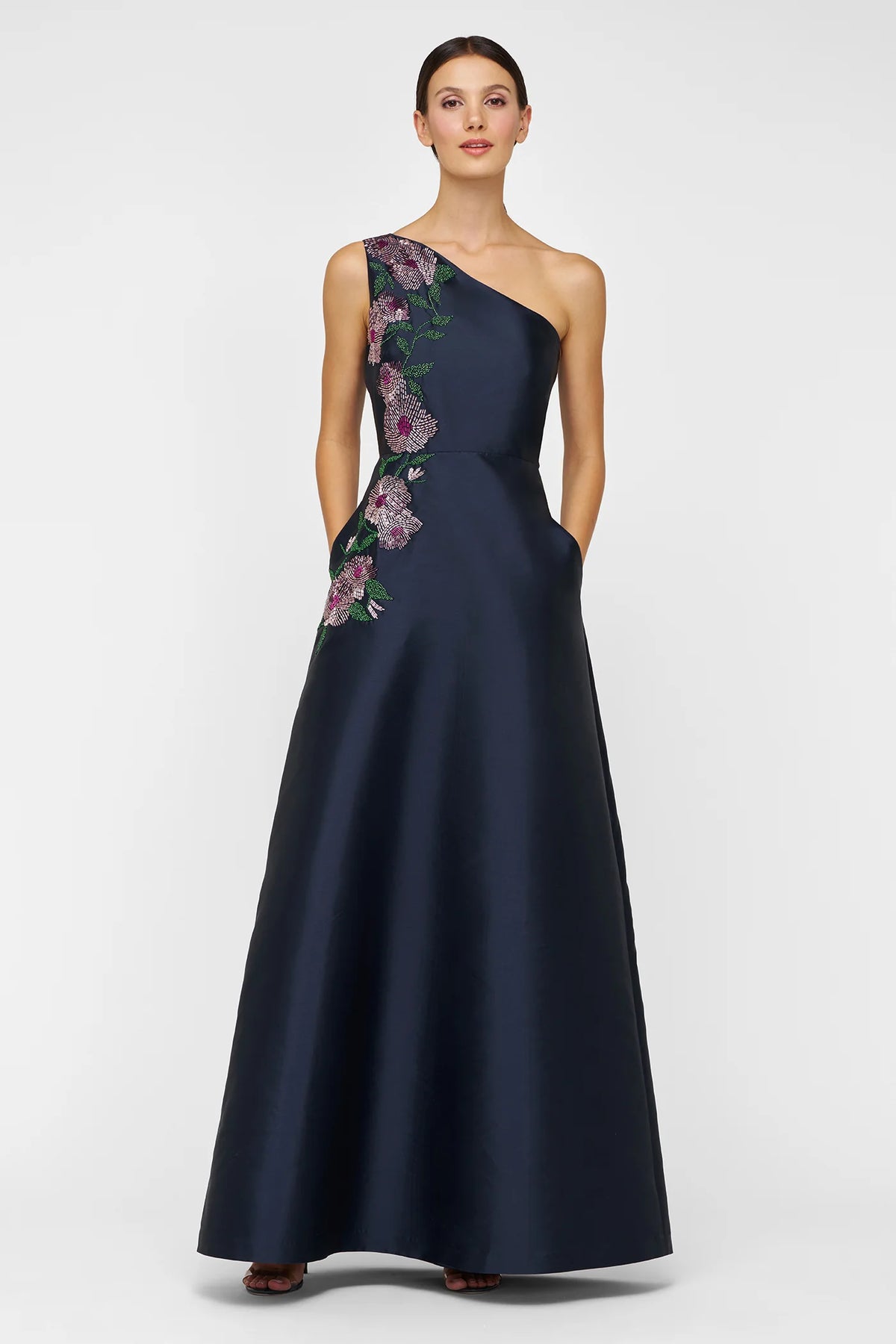 A model showcases a stunning navy one-shoulder gown featuring intricate floral beadwork along the neckline and side.