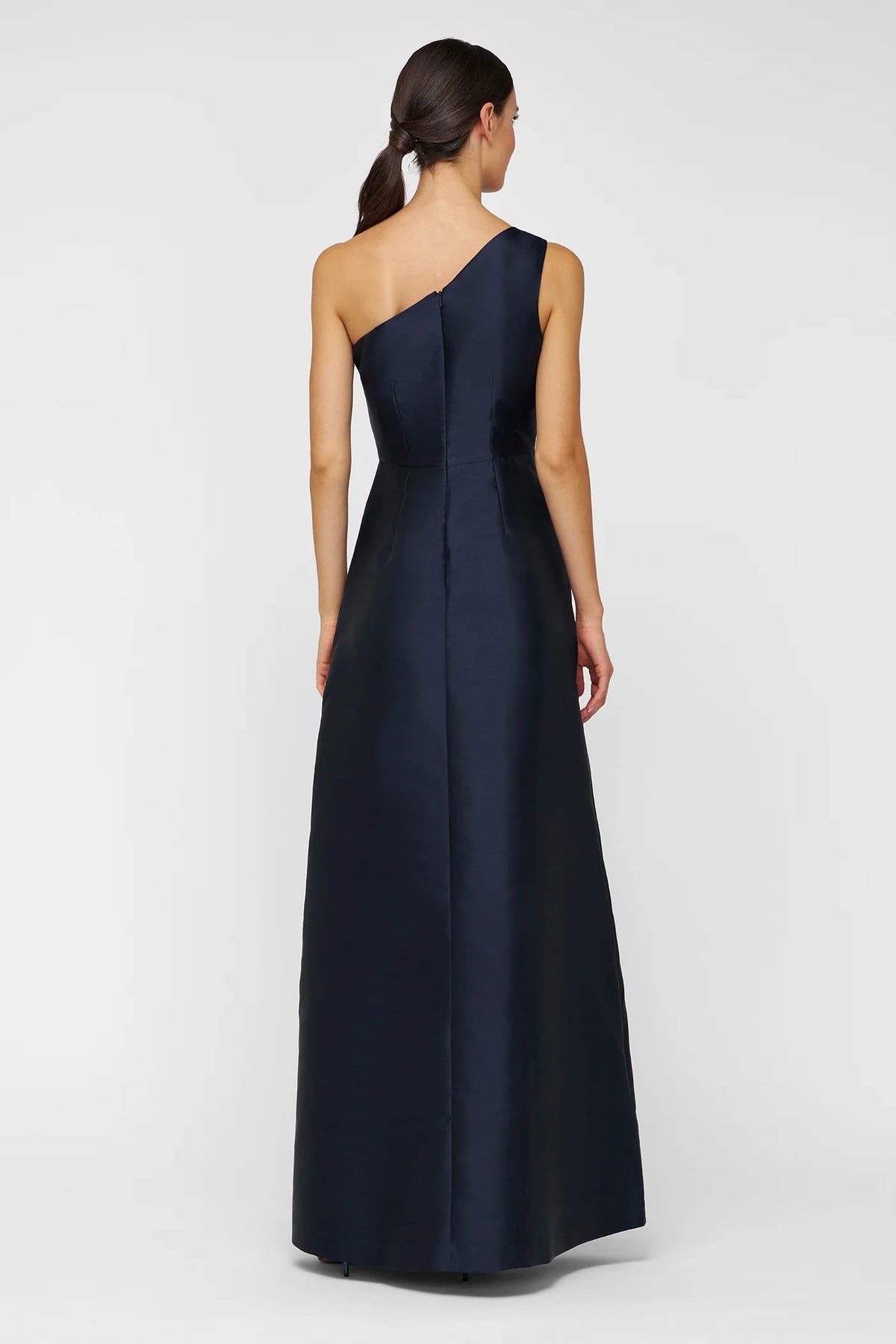 A model showcases a sleek, navy one-shoulder gown with a fitted bodice and flowing A-line skirt, perfect for formal events like black tie weddings or galas. This elegant dress is conveniently available near Apex, Cary, and Raleigh.