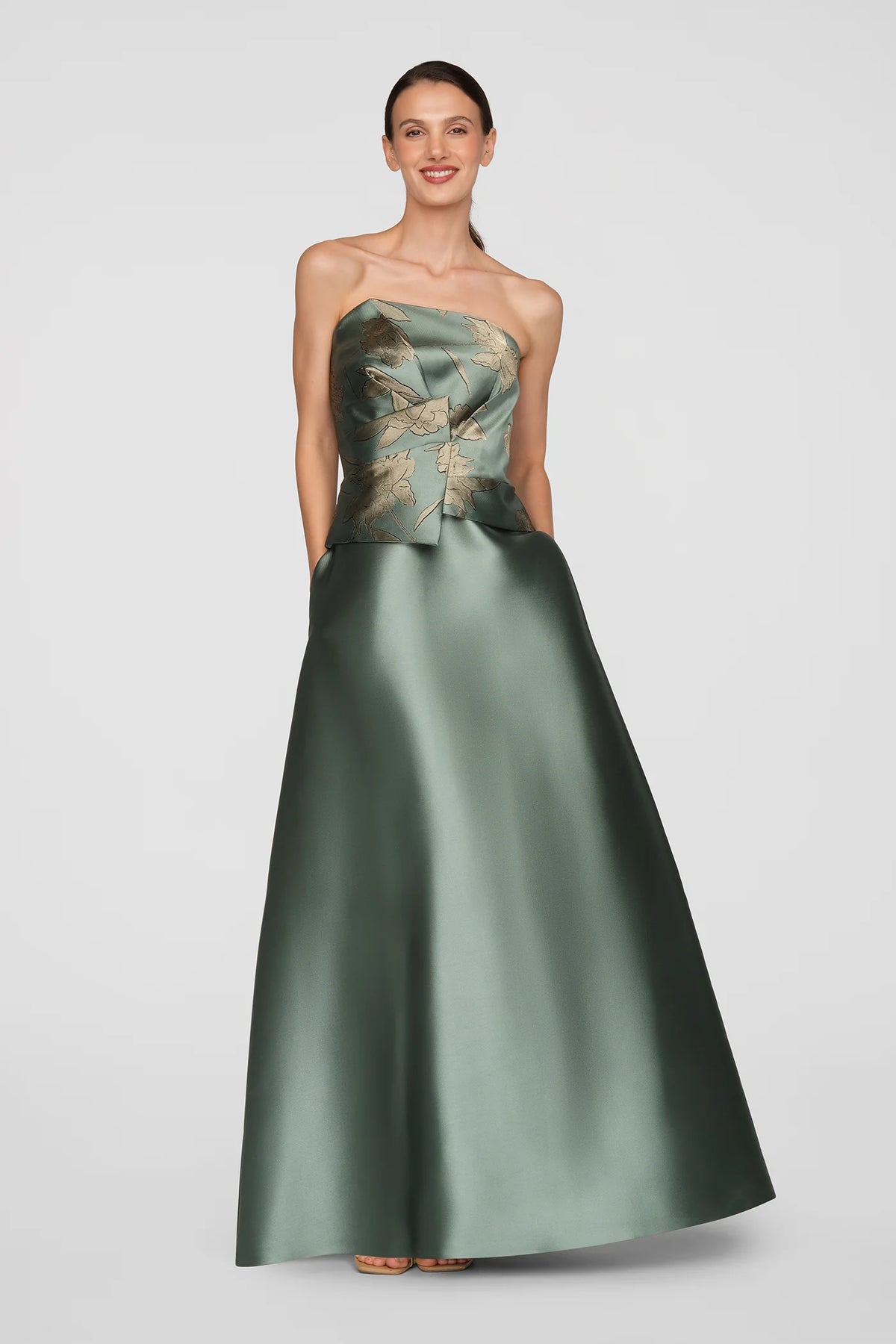 A model showcases a stunning soft sage ball gown with metallic jacquard folds and gold floral detailing, perfect for elegant occasions like mother of the bride, prom, or black tie weddings. Available for pre-order near Apex, Cary & Raleigh.