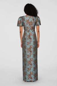 A model showcases the Esmerelda Dress from the back, featuring a vintage floral pattern in cool blue shades on a floor-length gown. Ideal for elegant occasions like black tie weddings or galas in the Apex, Cary, and Raleigh areas.