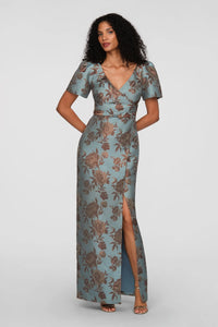 A model showcases a vintage floral jacquard gown featuring a faux-surplice bodice and a front slit, perfect for formal events like black tie weddings or galas. This elegant dress is conveniently located near Apex, Cary, and Raleigh.