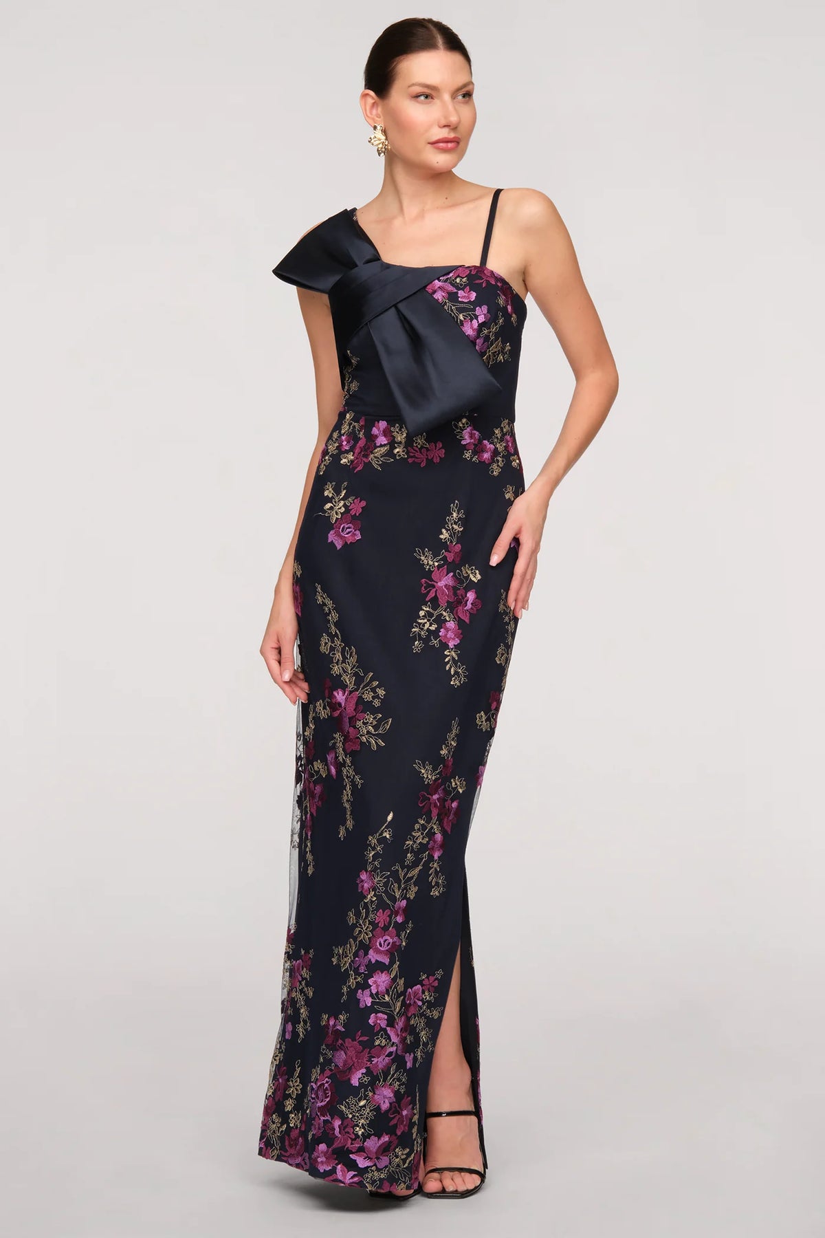 A model showcases a stunning navy gown adorned with vibrant floral patterns and an oversized bow on one shoulder.