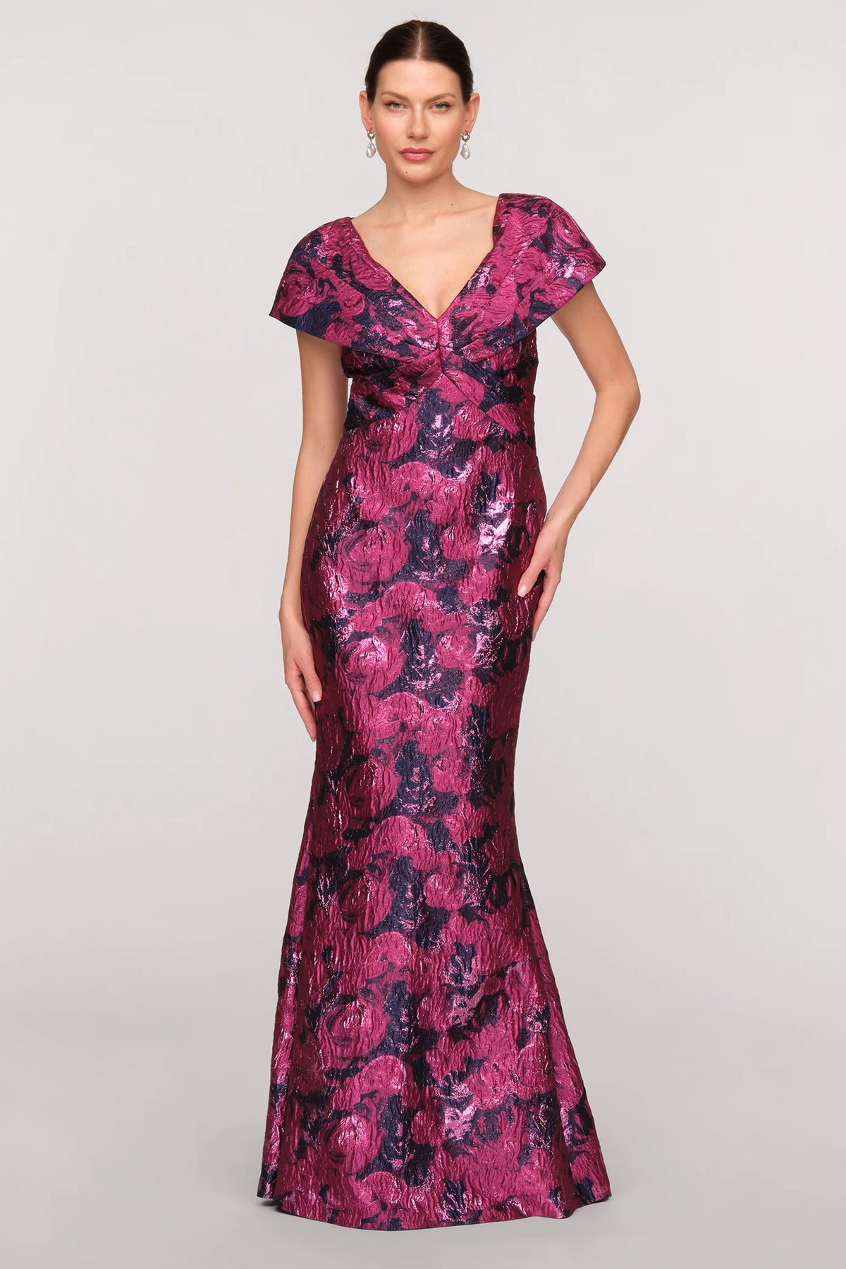 A model showcases a stunning mermaid gown featuring a vibrant floral jacquard in shades of purple and pink, complete with a V-neck and dramatic cape sleeves.