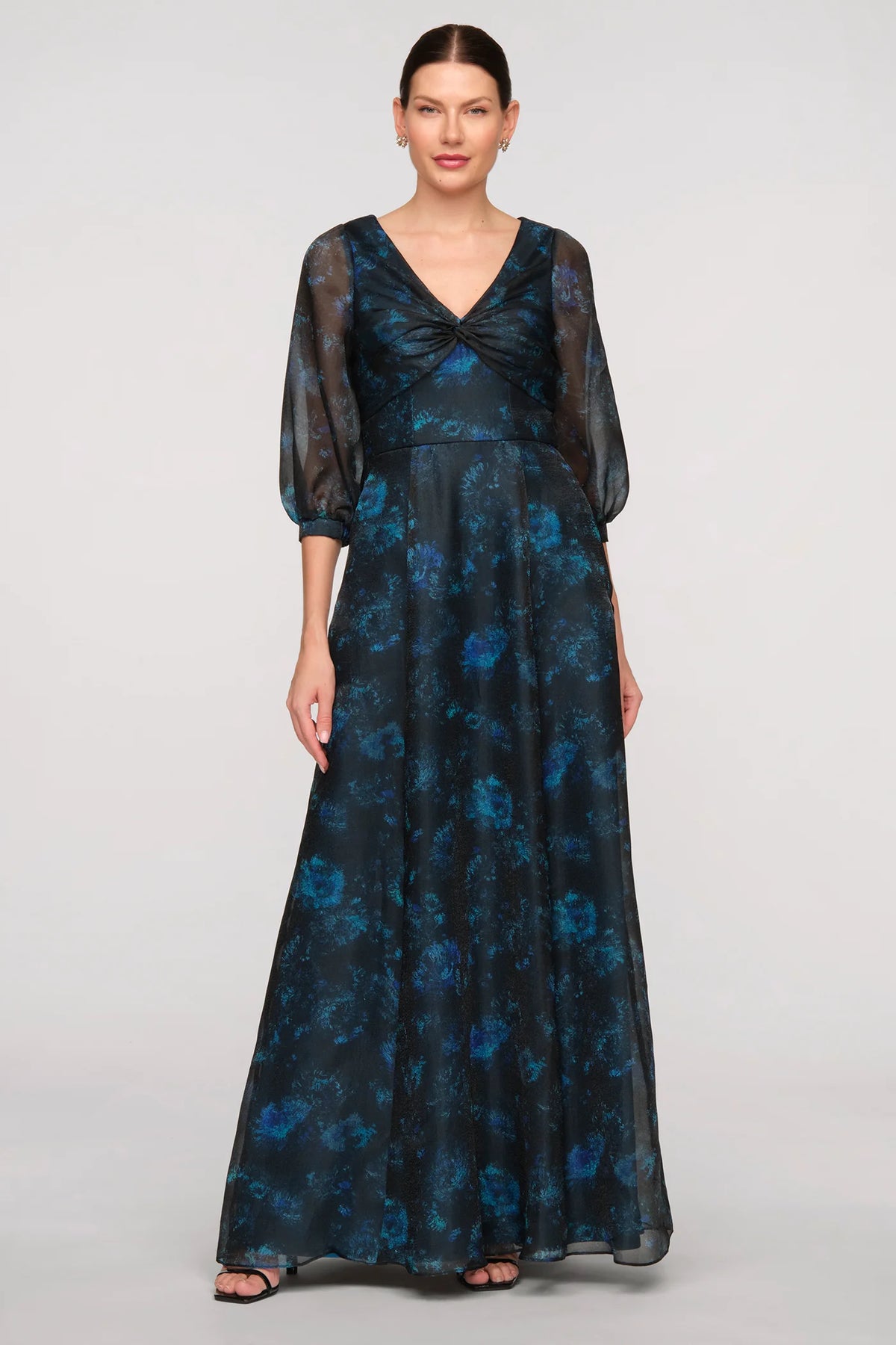 A model wears a floor-length, teal and blue printed organza gown featuring a twisted V-neck bodice and sheer balloon sleeves.