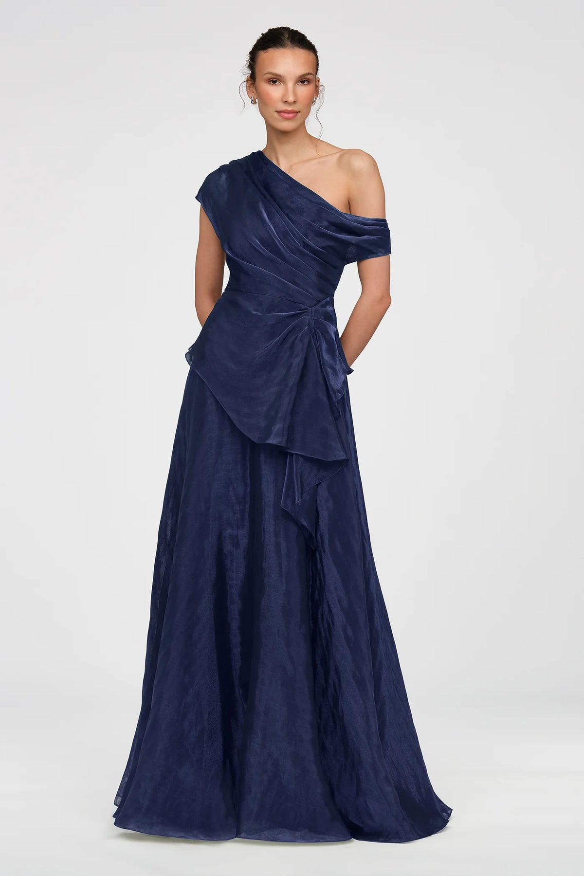 A model stands elegantly in a floor-length, one-shoulder navy gown featuring a soft peplum overlay and sculptural ruching.