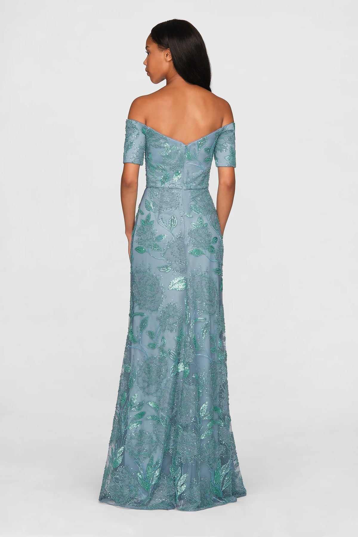 A woman is seen from the back wearing a stunning teal blue off-shoulder gown adorned with intricate beadwork and floral designs.