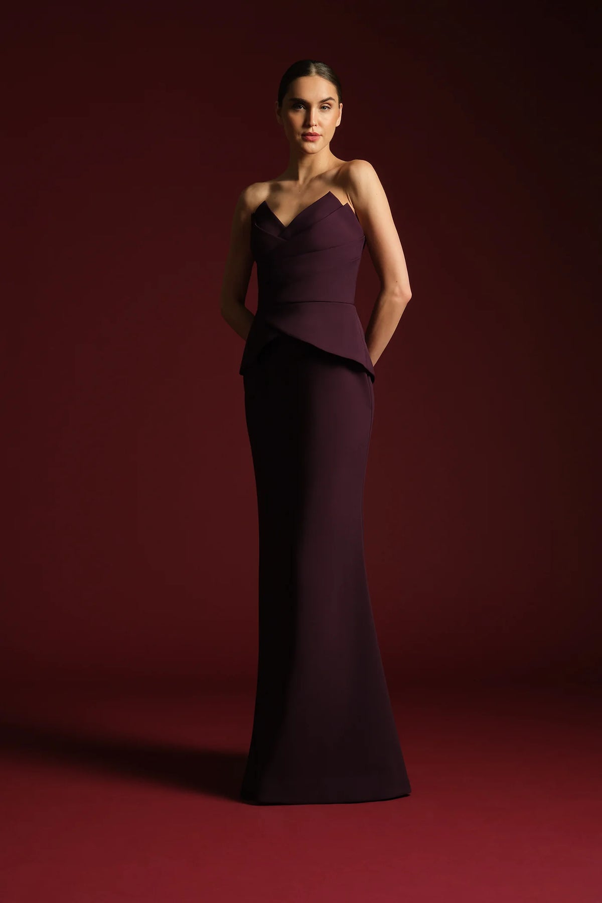 A model showcases a sleek, strapless, floor-length gown in a rich plum color, featuring a fitted bodice and elegant peplum detail.