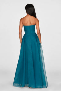 A model showcases a stunning strapless draped gown in a rich teal color, featuring a fitted bodice and a flowing skirt with sheer layers.
