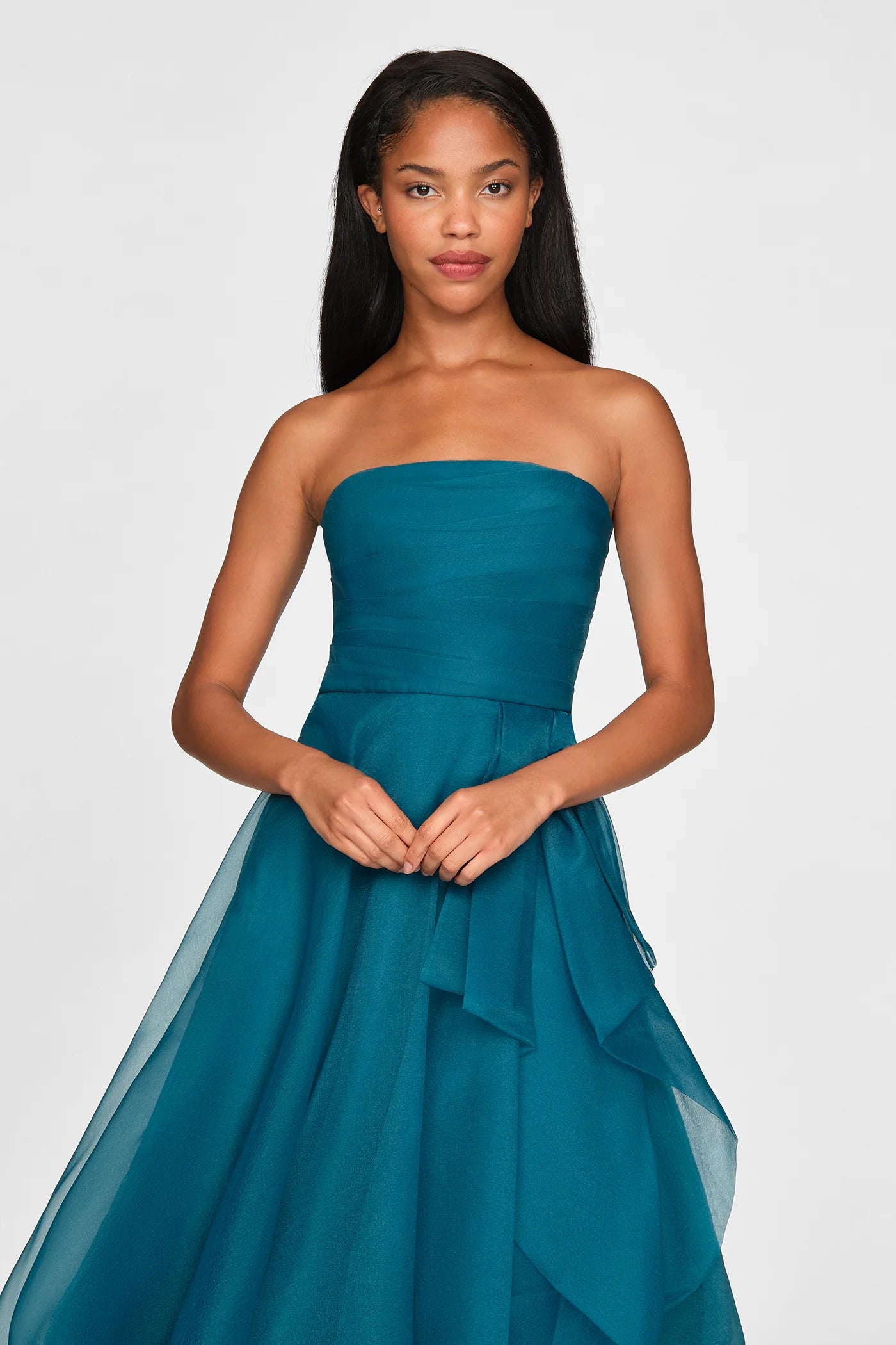 A model poses in a strapless teal gown featuring a draped design and flowing layers. This elegant dress is perfect for special occasions like proms or galas and is conveniently located near Apex, Cary, and Raleigh.