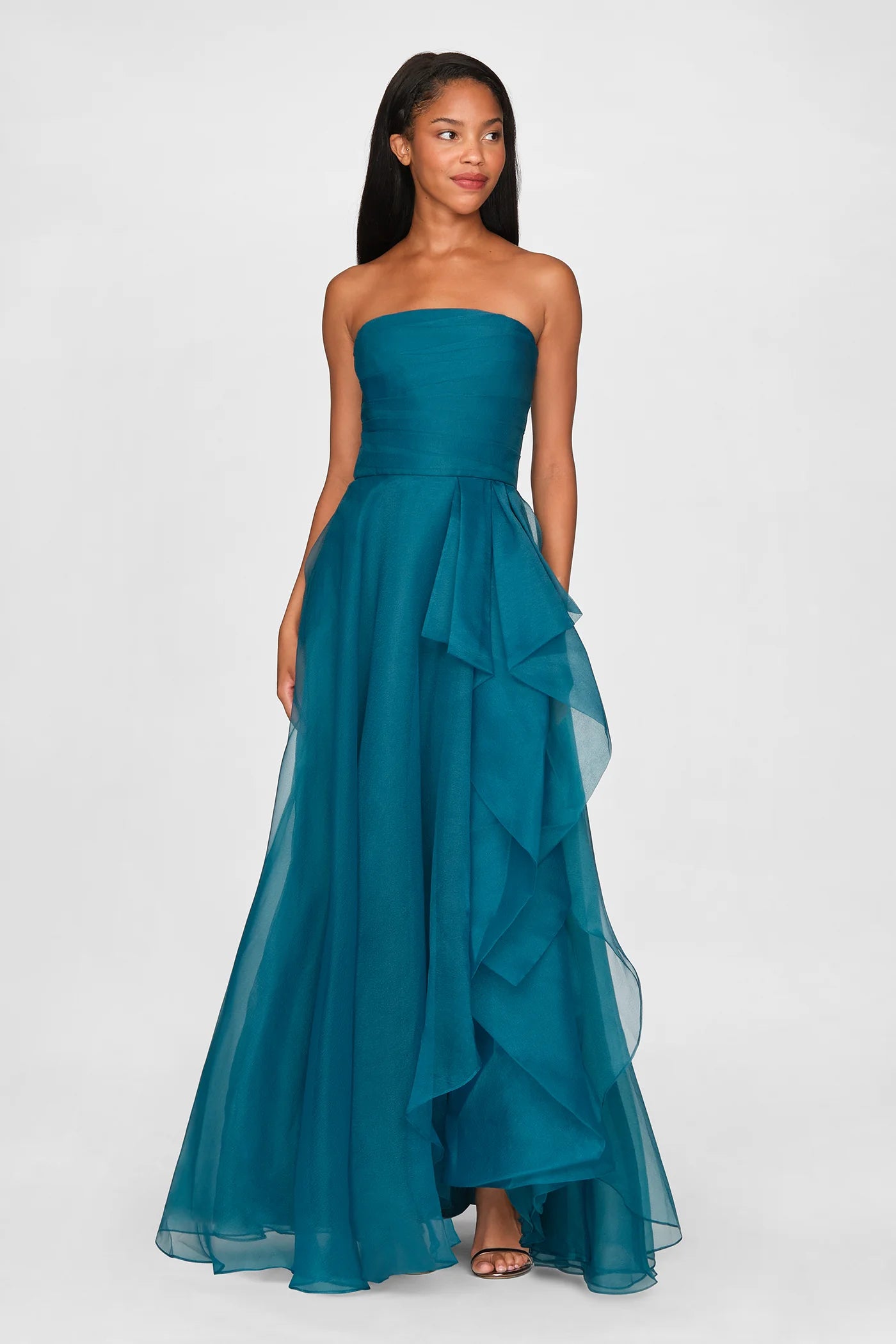 A model showcases a stunning strapless draped gown in a rich teal color, featuring soft, flowing layers and a side ruffle.