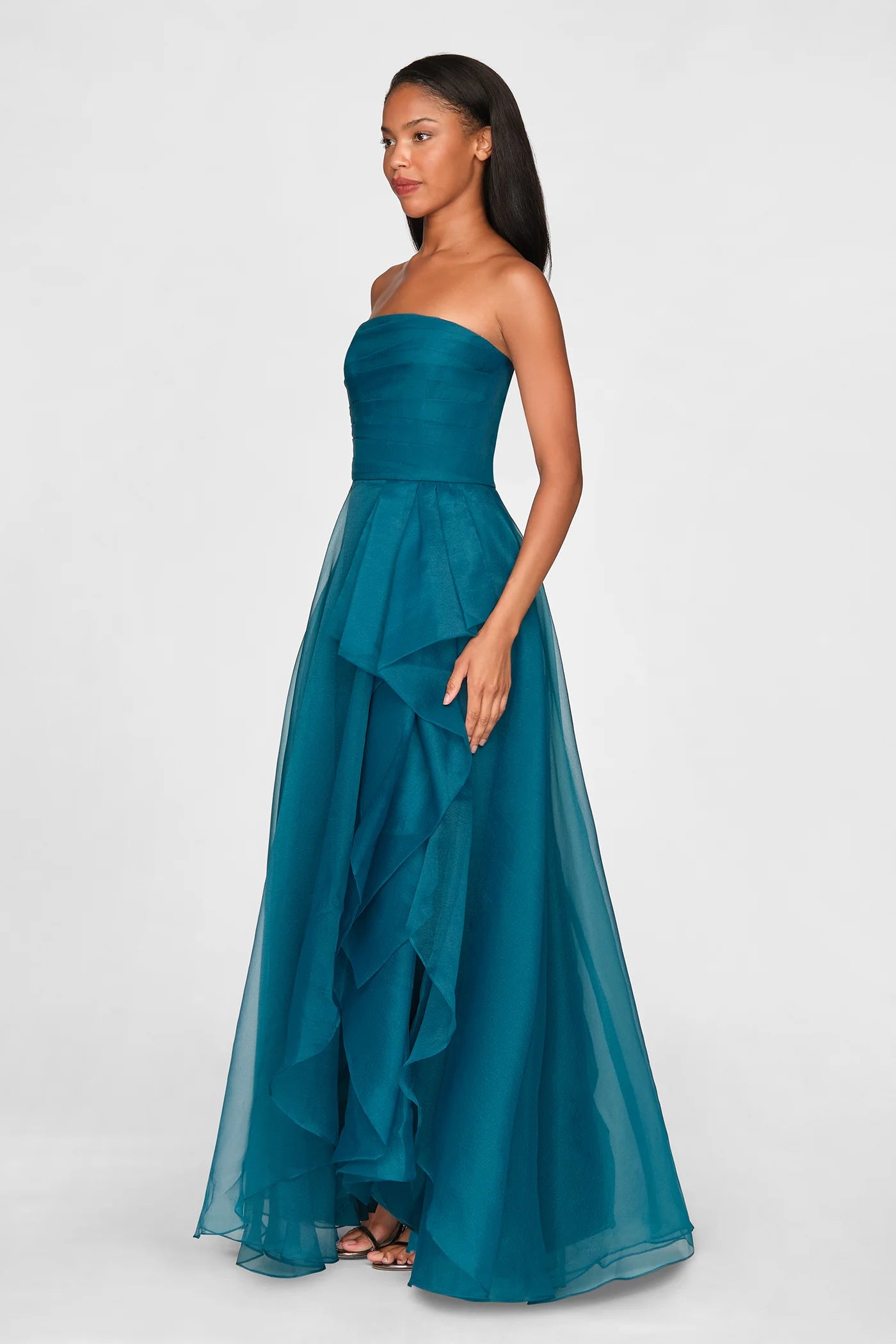 A model showcases a stunning strapless draped gown in a rich teal color, featuring layered ruffles and a flowing silhouette.