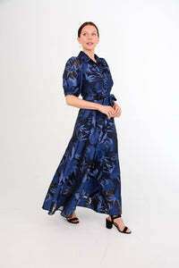 A model is wearing a long, navy blue maxi dress with bold floral patterns and puff sleeves, styled with a belt at the waist.
