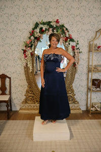 A woman poses confidently in a navy gown with a strapless design and intricate embellishments, standing in front of an ornate mirror adorned with flowers.