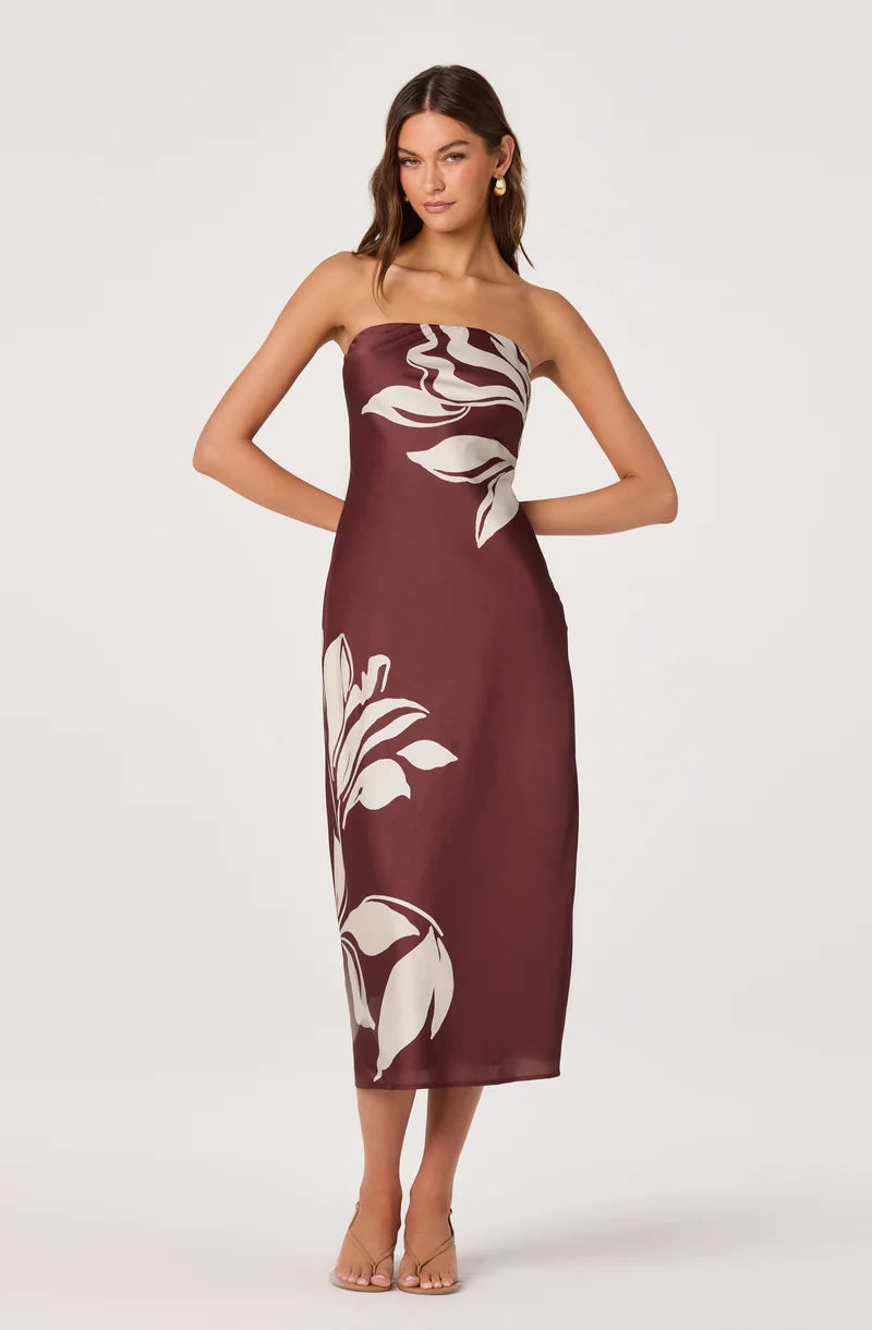 A model showcases a strapless, burgundy maxi dress adorned with large white floral patterns. This elegant gown is perfect for formal occasions such as weddings, galas, or proms, and is conveniently available near Apex, Cary, and Raleigh.