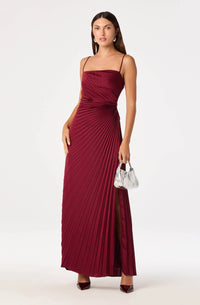 A model poses in a rich burgundy satin pleated maxi dress with adjustable spaghetti straps and a side leg slit, ideal for galas or black-tie events. This stunning gown is conveniently located near Apex, Cary, and Raleigh.